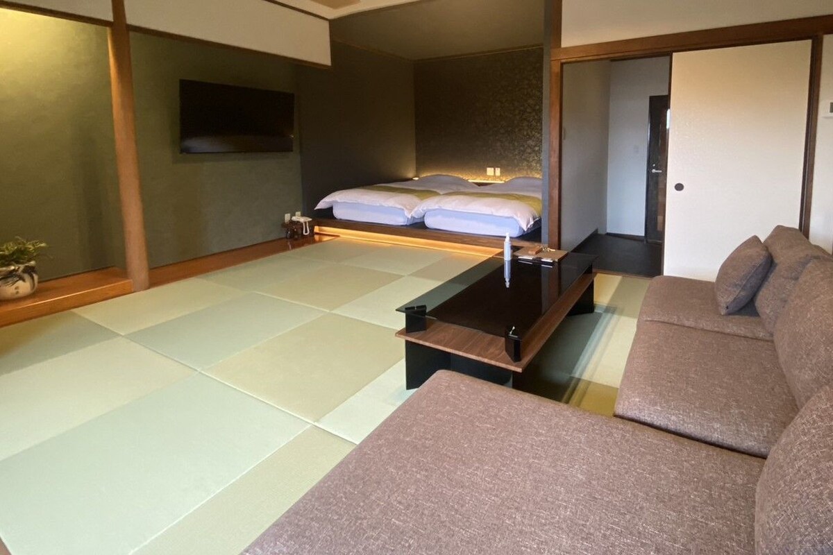 Une pièce de style japonais traditionnel, avec un sol recouvert de tatamis, un lit bas en bois, un canapé et un téléviseur fixé au mur.