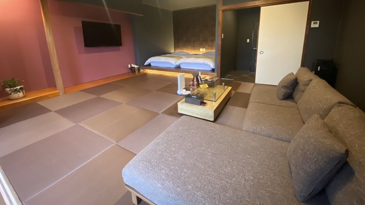 A modern hotel room with a large sofa, a small table with a tray, a bed with white linens, and a television mounted on the wall.