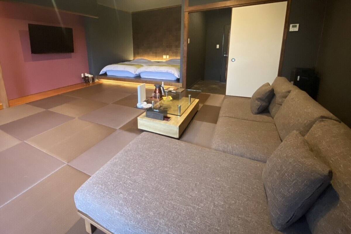 Quarto de hotel moderno com um sofá grande, uma mesinha com bandeja, cama com roupa de cama branca e uma televisão fixada na parede.