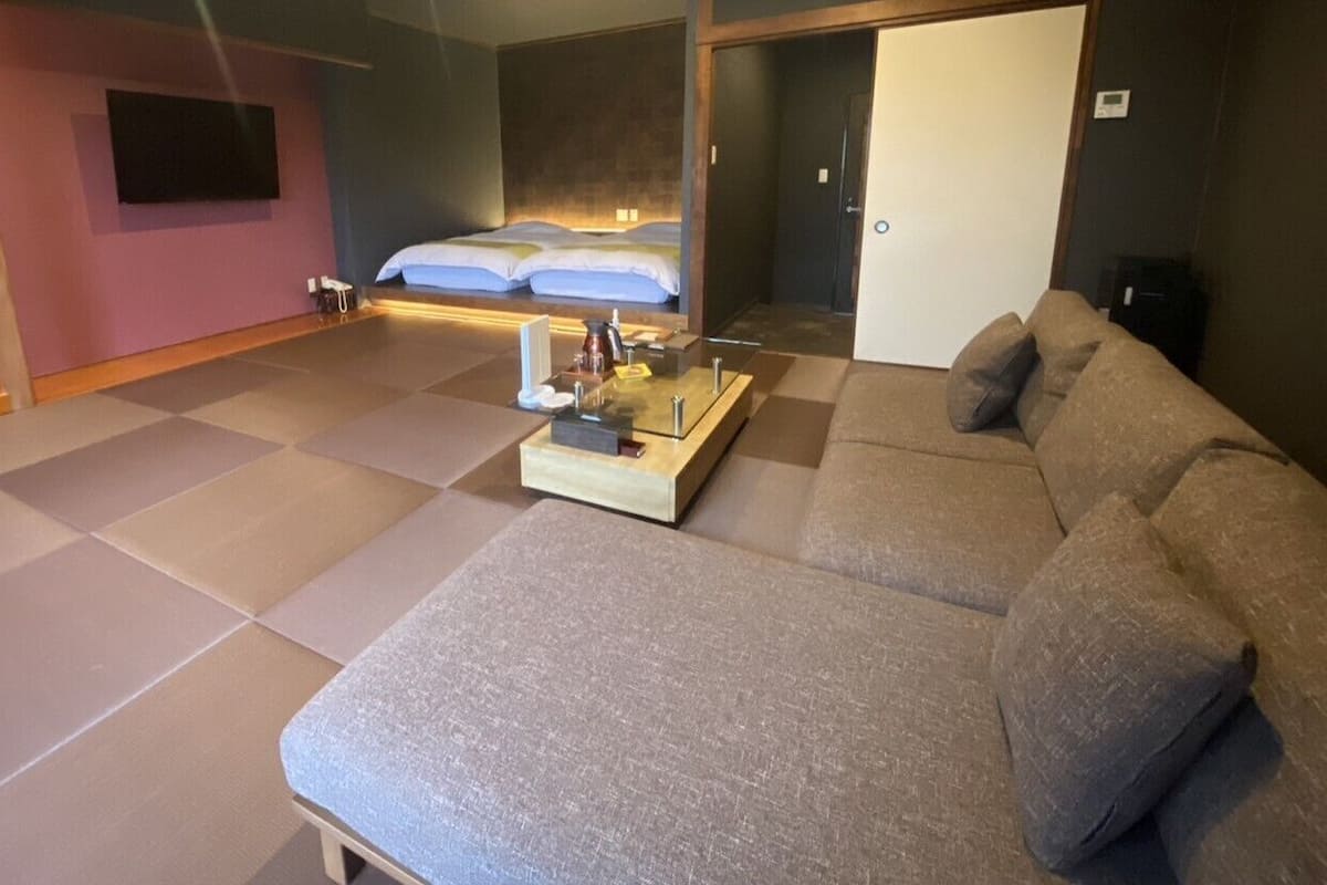 A modern hotel room with a large sofa, a small table with a tray, a bed with white linens, and a television mounted on the wall.