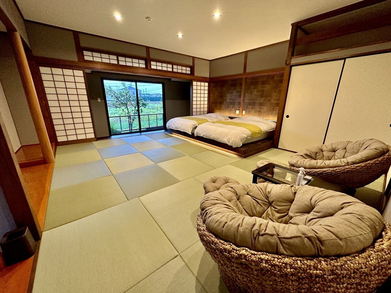 A tatami mat room with a bed, two tatami chairs, and a sliding door leading to a balcony.