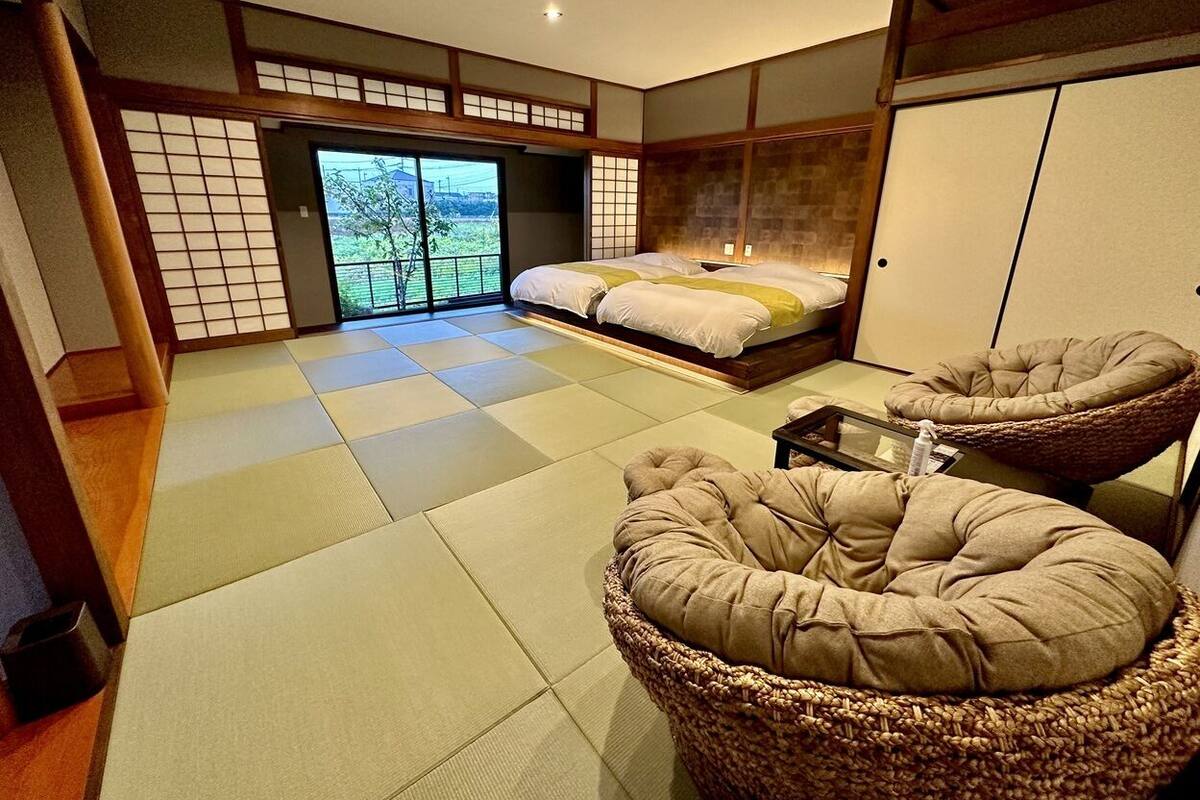 Um quarto com esteiras de tatame, uma cama, duas cadeiras de tatame e uma porta deslizante para uma varanda.