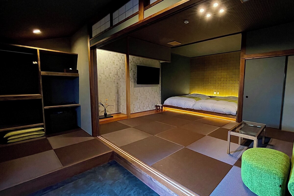 Une chambre d’hôtel moderne, équipée d’un lit, d’une petite table, d’une chaise verte, d’un dressing et offrant une vue sur la ville.