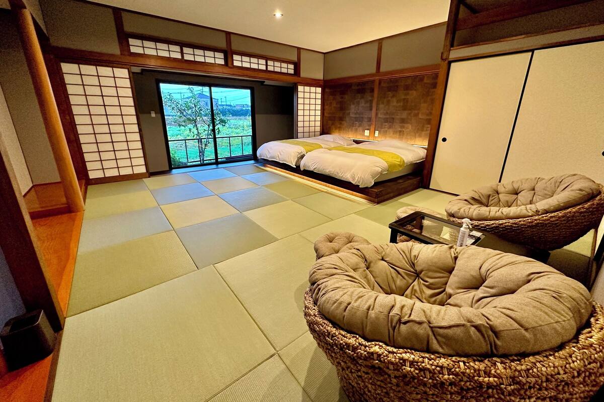 Um quarto em estilo japonês tradicional com tatames, uma cama com roupa de cama branca, um futon e uma cadeira de fibra vegetal.