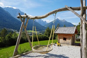 Un columpio de madera con asiento de mimbre, una pequeña cabaña de madera y un tractor en un entorno rural con montañas al fondo.