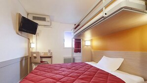 Ein Einzelbett mit roter Bettdecke, ein Nachttisch, ein an der Wand montierter Fernseher und ein Fenster mit Vorhängen.