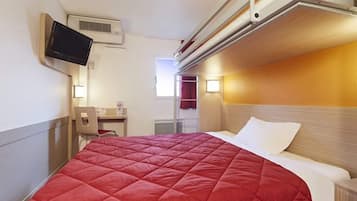 A single bed with a red quilt, a bedside table, a TV mounted on the wall, and a window with curtains.