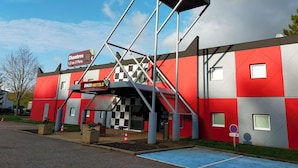 A hotel with a red and grey exterior, a signboard for ENZO HOTELS, and a ramp leading to the entrance. - Hotel Lemon (Coudes)