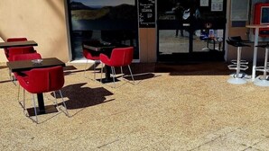 An outdoor seating area with tables and red chairs, a vending machine, and a view of the interior through a glass door. - Hotel Lemon (Coudes)