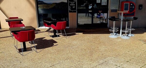 An outdoor seating area with tables and red chairs, a vending machine, and a view of the interior through a glass door. - Hotel Lemon (Coudes)