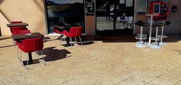 Un área con mesas y sillas rojas para sentarse al aire libre, una máquina expendedora y vista del interior a través de una puerta de vidrio.