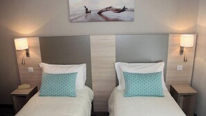 Ein Hotelzimmer mit zwei Betten, jeweils mit weißer Bettdecke und Kissen mit blaugrünem Muster, einem großen Wandbild, das eine Naturlandschaft zeigt, und Nachttischlampen an beiden Seiten der Betten.