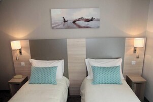 Habitación de hotel con dos camas, cada una con una colcha blanca y almohadas con estampado azul verdoso, un cuadro grande colgado en la pared que representa una escena natural y lámparas de noche a ambos lados de las camas.