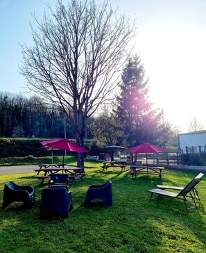Een buitenzitplaats met picknicktafels en stoelen, omgeven door bomen en een gebouw op de achtergrond.