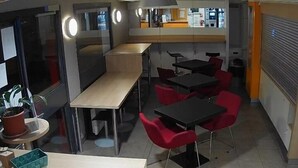 Un intérieur moderne avec une longue table et plusieurs chaises rouges, une petite table ronde avec un pot de plante, et un comptoir avec une caisse enregistreuse.