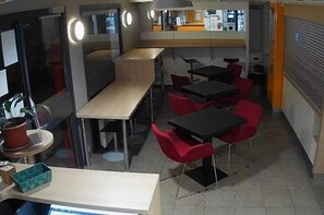 A modern interior with a long table and several red chairs, a small round table with a potted plant, and a counter area with a cash register. - Hotel Lemon (Coudes)