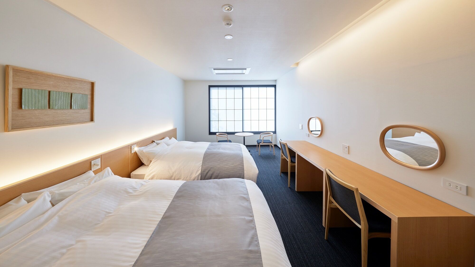 Ein Hotelzimmer mit zwei Betten, einem Schreibtisch, einem Stuhl, einem runden Spiegel und einem Fenster.