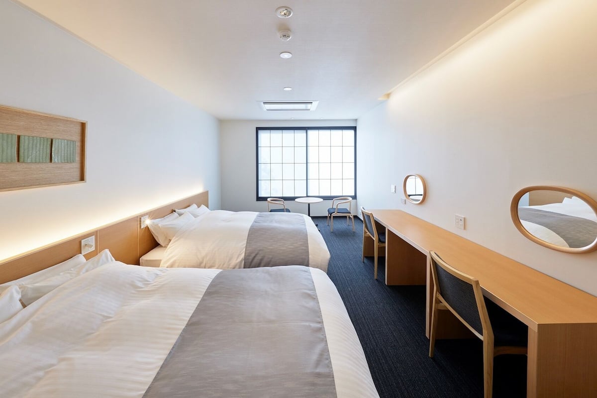 Une chambre d’hôtel avec deux lits, un bureau, une chaise, un miroir rond et une fenêtre.