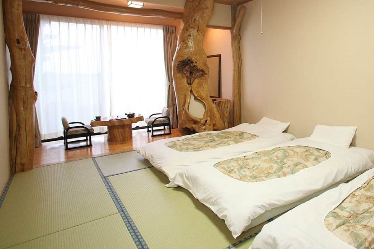 Une pièce avec deux lits, un sol recouvert de tatamis, un mur décoré avec un tronc d’arbre en bois, une table à manger avec des chaises et une grande fenêtre avec des rideaux.