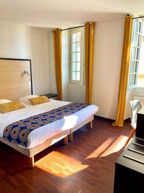 A hotel room with a bed, wooden floor, a desk, and curtains.