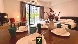 Hotelkamer met een ontbijt gedekte eettafel, een bed met groene hoofdbord, een vaas met bloemen, en uitzicht buiten door grote ramen.