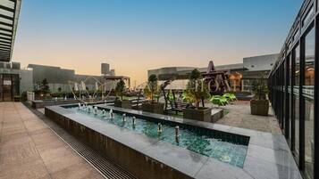 A rooftop pool area with a long rectangular pool, surrounded by potted plants and seating areas.
