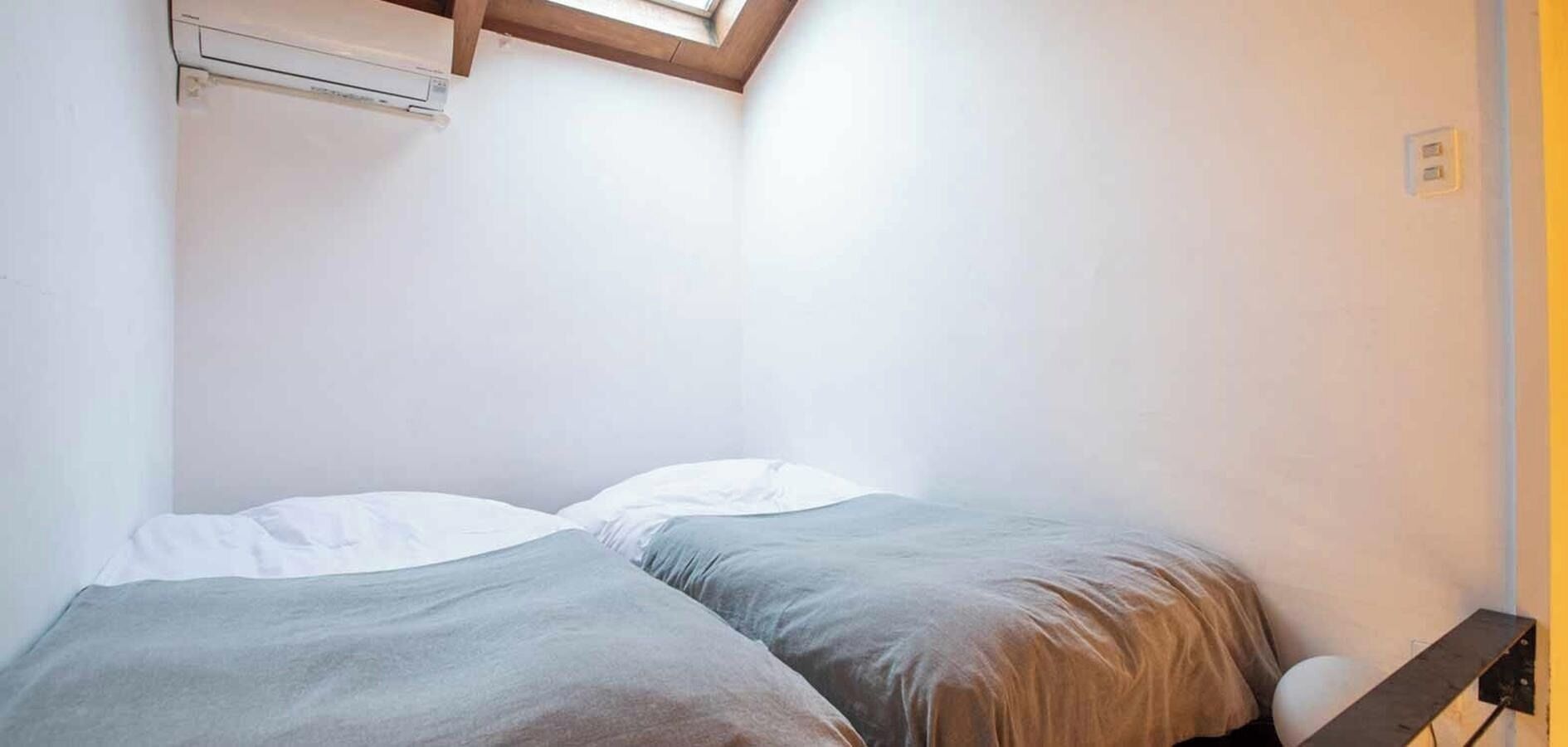 A small, single-bed room with a slanted ceiling, a wooden headboard, and a wall-mounted air conditioning unit.