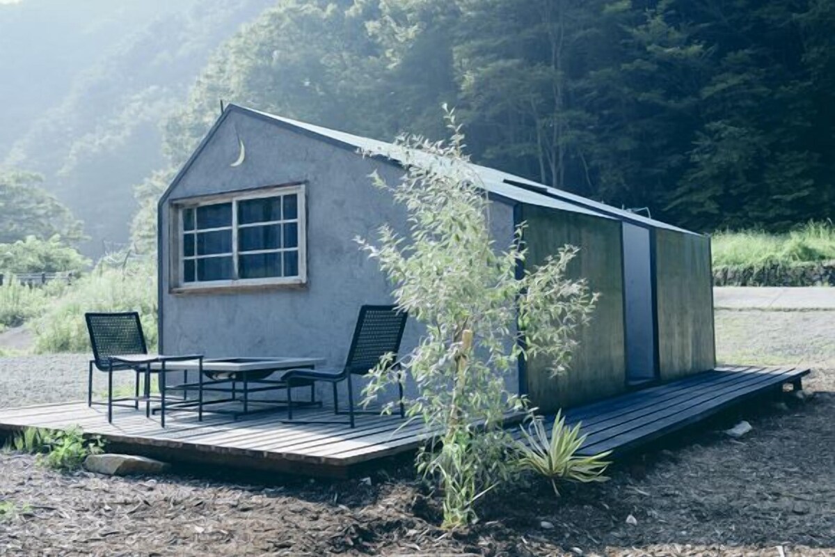 Una cabaña pequeña con terraza de madera, dos sillas y una mesa.