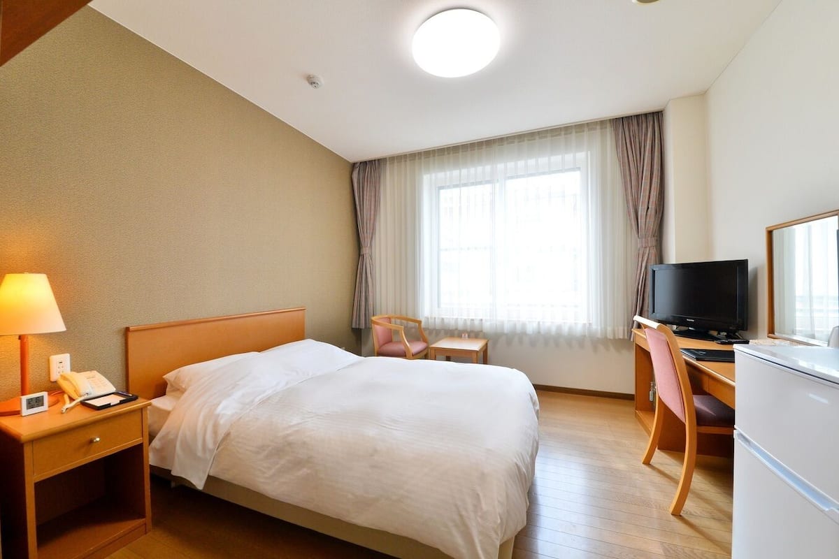 A hotel room with a bed, bedside table with a lamp, a desk with a chair, a television, and a window with curtains.