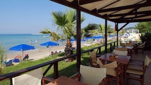 A covered outdoor seating area with tables and chairs, overlooking a beach with umbrellas and palm trees. - Alykanas Beach Grand Hotel by Zante Plaza (Zakynthos)
