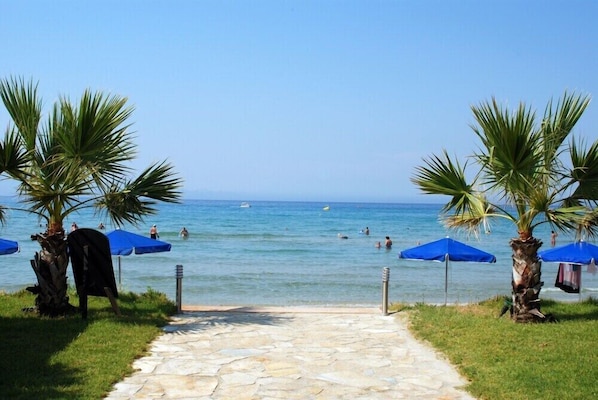 A stone pathway leading to a beach with palm trees and blue umbrellas.