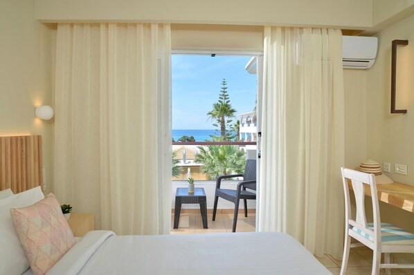 A hotel room with a bed, a small table, a chair, a desk, and a view of the sea and palm trees.