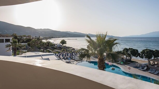 A pool area with lounge chairs and a palm tree, overlooking a beach and mountains. - Alykanas Beach Grand Hotel by Zante Plaza (Zakynthos)