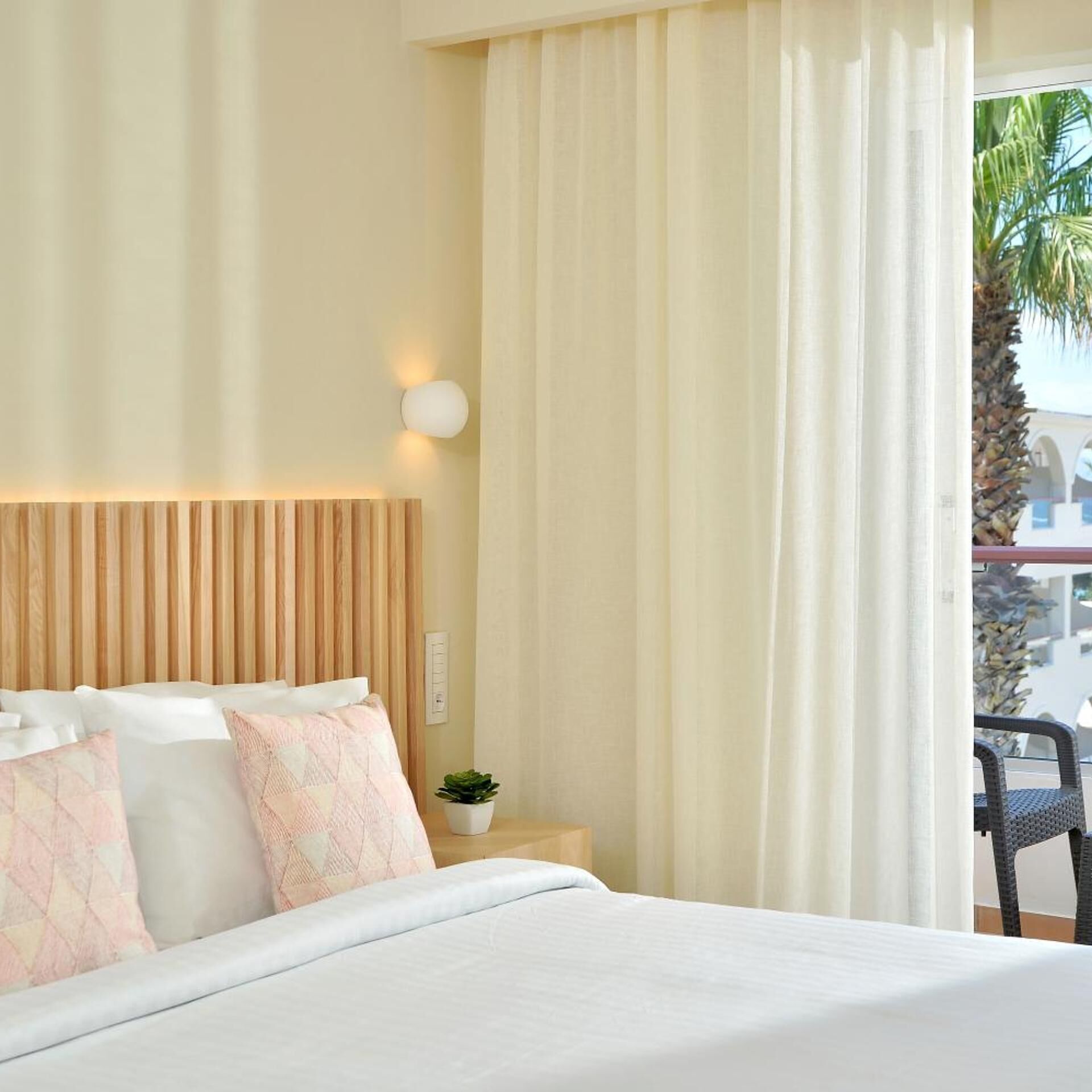 Habitación de hotel con cama, mesita de noche con lámpara, planta en maceta y vista a palmeras desde el exterior.