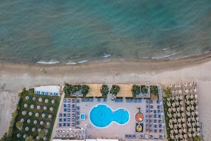 An aerial view of a beachside resort with a swimming pool, sun loungers, umbrellas, and a beach area.