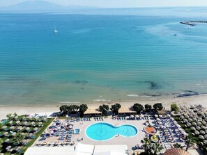 Eine Vogelperspektive zeigt ein Resort mit einem zentralen Infinity-Pool, Liegebereichen am Strand und Blick auf das Meer.