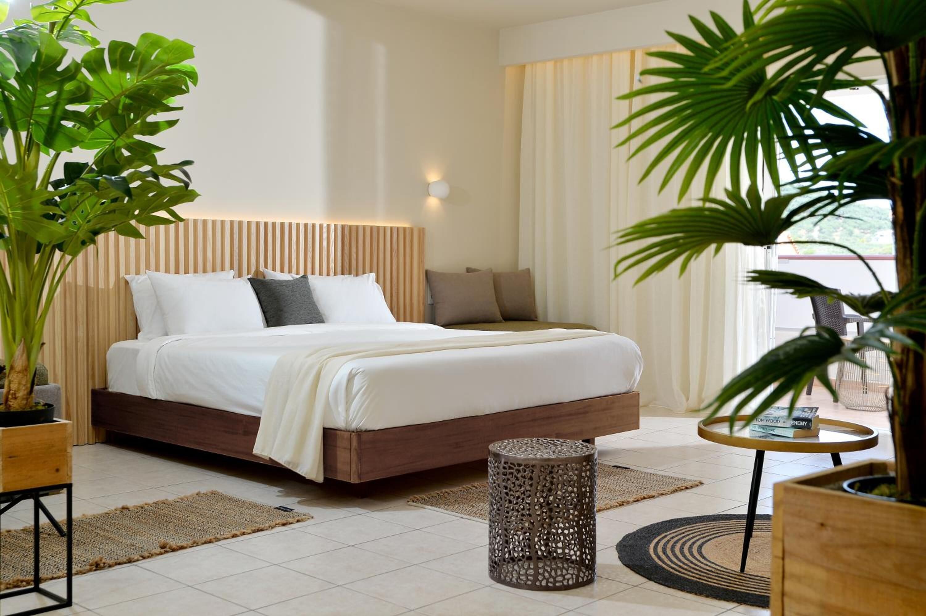Una habitación de hotel con cama, mesita de noche, planta en maceta y ventana con cortinas.