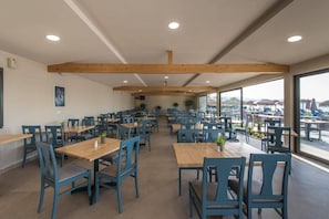 A spacious dining area with wooden tables and blue chairs, large windows offering a view of the beach.
