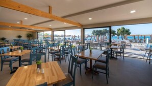 A spacious dining area with wooden tables and chairs, large windows offering a view of the sea and buildings. - Alykanas Beach Grand Hotel by Zante Plaza (Zakynthos)