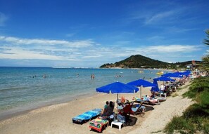Uma praia com espreguiçadeiras e guarda-sóis, céu claro e paisagem montanhosa ao fundo.