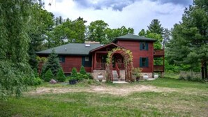 A two-story house with a red exterior, a front porch, and a garden with various plants and a small tree. - Secluded 4-bedroom log cabin surrounded by beautiful forest (New Lisbon)