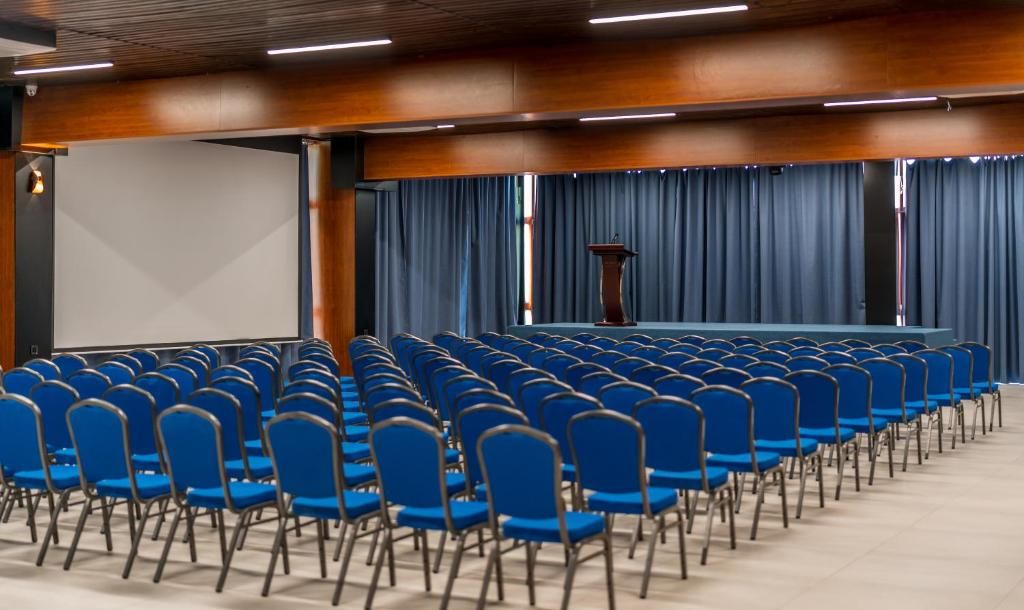 Uma sala de conferências com fileiras de cadeiras azuis, um púlpito e uma tela grande.