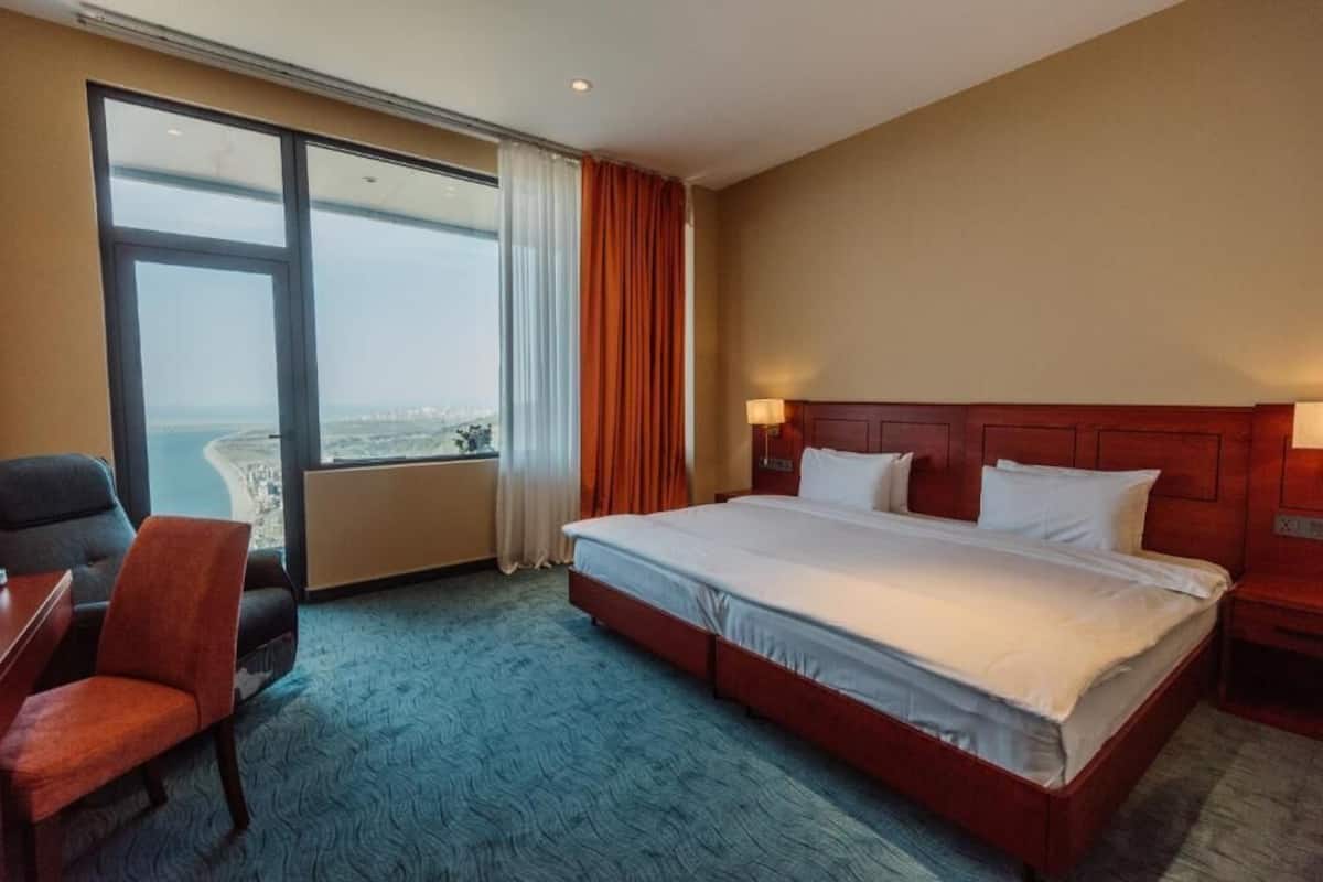 A hotel room with a large bed, a desk, a chair, and a view of the city through a large window.