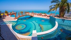 A pool with a circular design, surrounded by lounge chairs and a palm tree, overlooking a clear blue sea.