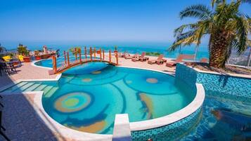 A pool with a circular design, surrounded by lounge chairs and a palm tree, overlooking a clear blue sea.
