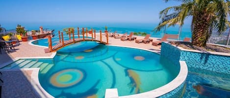 A pool with a circular design, surrounded by lounge chairs and a palm tree, overlooking a clear blue sea.