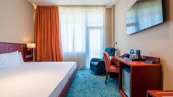 A hotel room with a bed, a desk with a chair, a television, and a window with curtains.