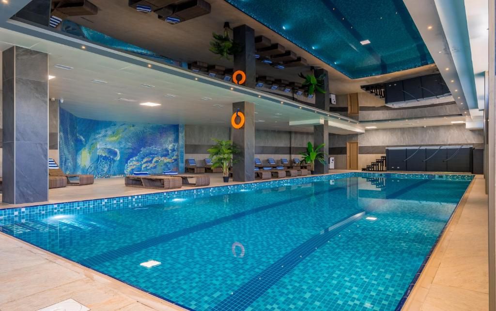 An indoor swimming pool with a tiled floor, a ceiling with recessed lighting, and a seating area with chairs and potted plants.