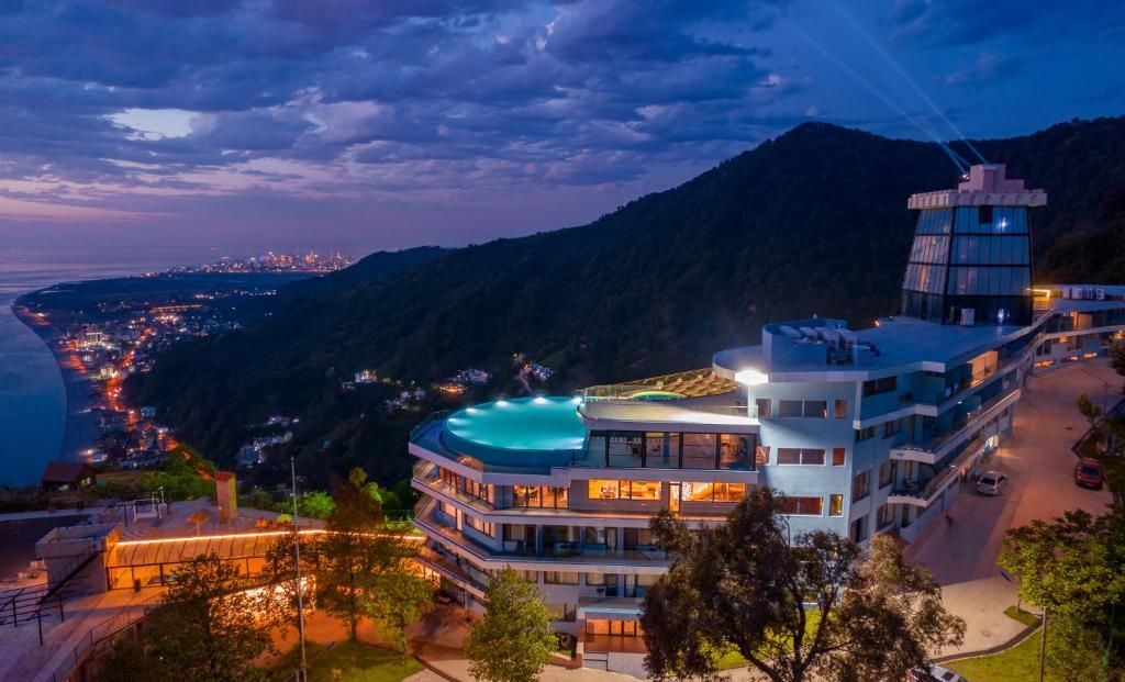 Un hotel de varios pisos con piscina en la azotea, rodeado de montañas y un paisaje urbano costero.