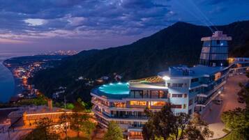 Un hotel de varios pisos con piscina en la azotea, rodeado de montañas y un paisaje urbano costero.